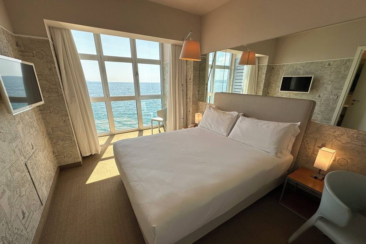 A hotel room with a large bed, a TV, a chair, and a view of the sea.