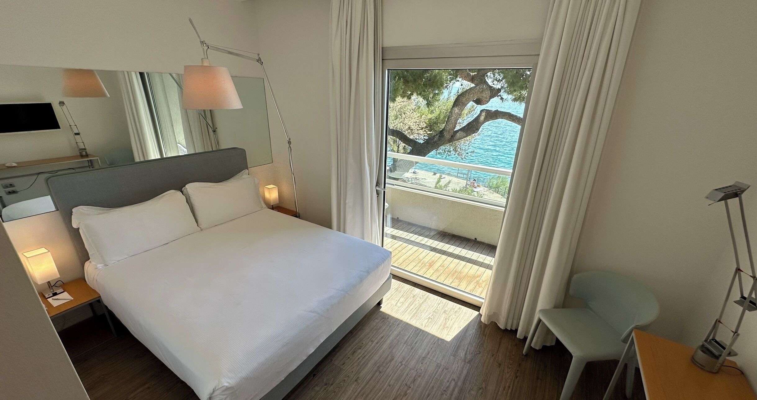 Habitación de hotel moderna con una cama grande, lámparas de noche, un televisor de pantalla plana y vista a una piscina y árboles.