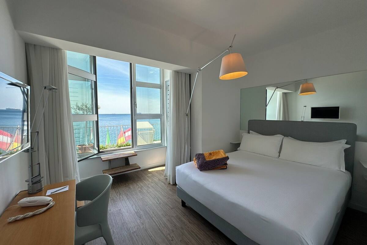 A modern hotel room with a large bed, a desk with a phone, and a view of the sea through large windows.