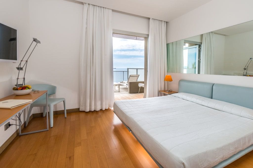 Una habitación de hotel moderna con una cama grande, un escritorio con una silla, una lámpara de escritorio y vista al mar a través de una puerta de balcón.