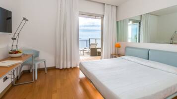 Una habitación de hotel moderna con una cama grande, un escritorio con una silla, una lámpara de escritorio y vista al mar a través de una puerta de balcón.