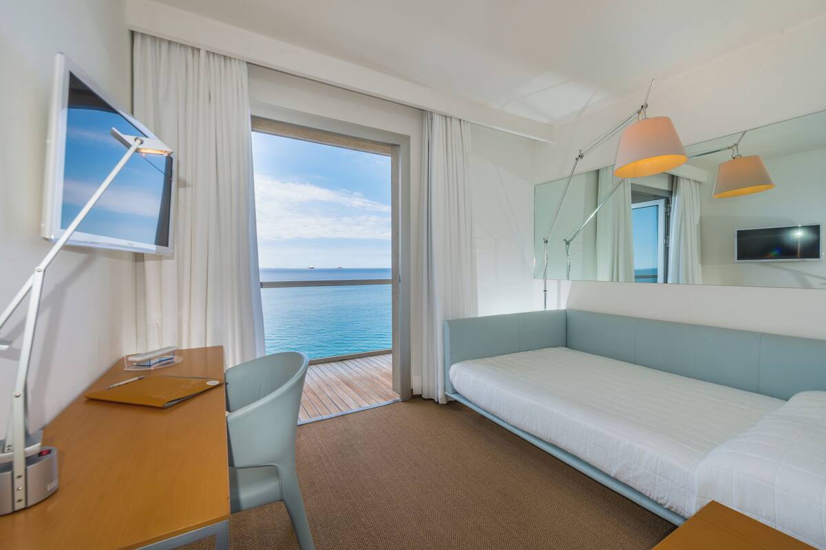 A hotel room with a bed, a desk, a chair, a TV, and a view of the sea.