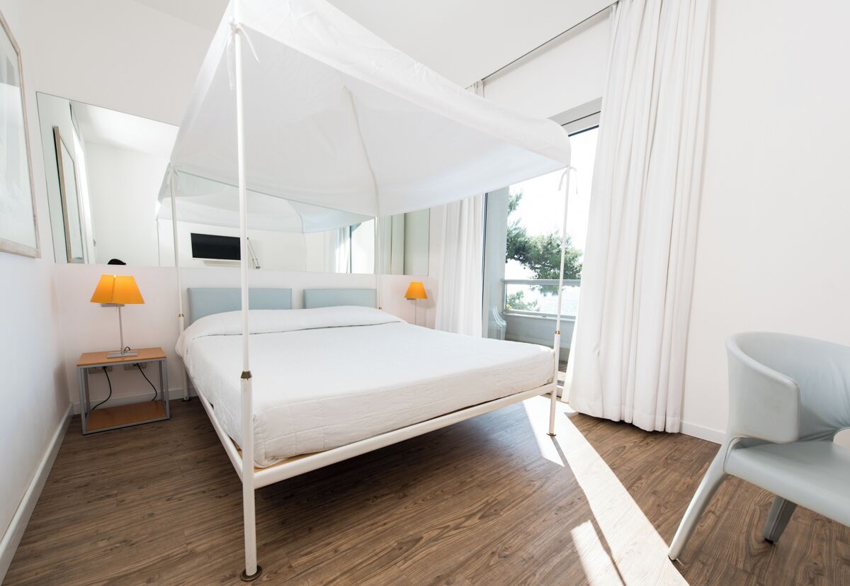 Un dormitorio moderno con cama con dosel, suelo de madera, una mesita de noche con lámpara, una silla y un gran ventanal con cortinas.