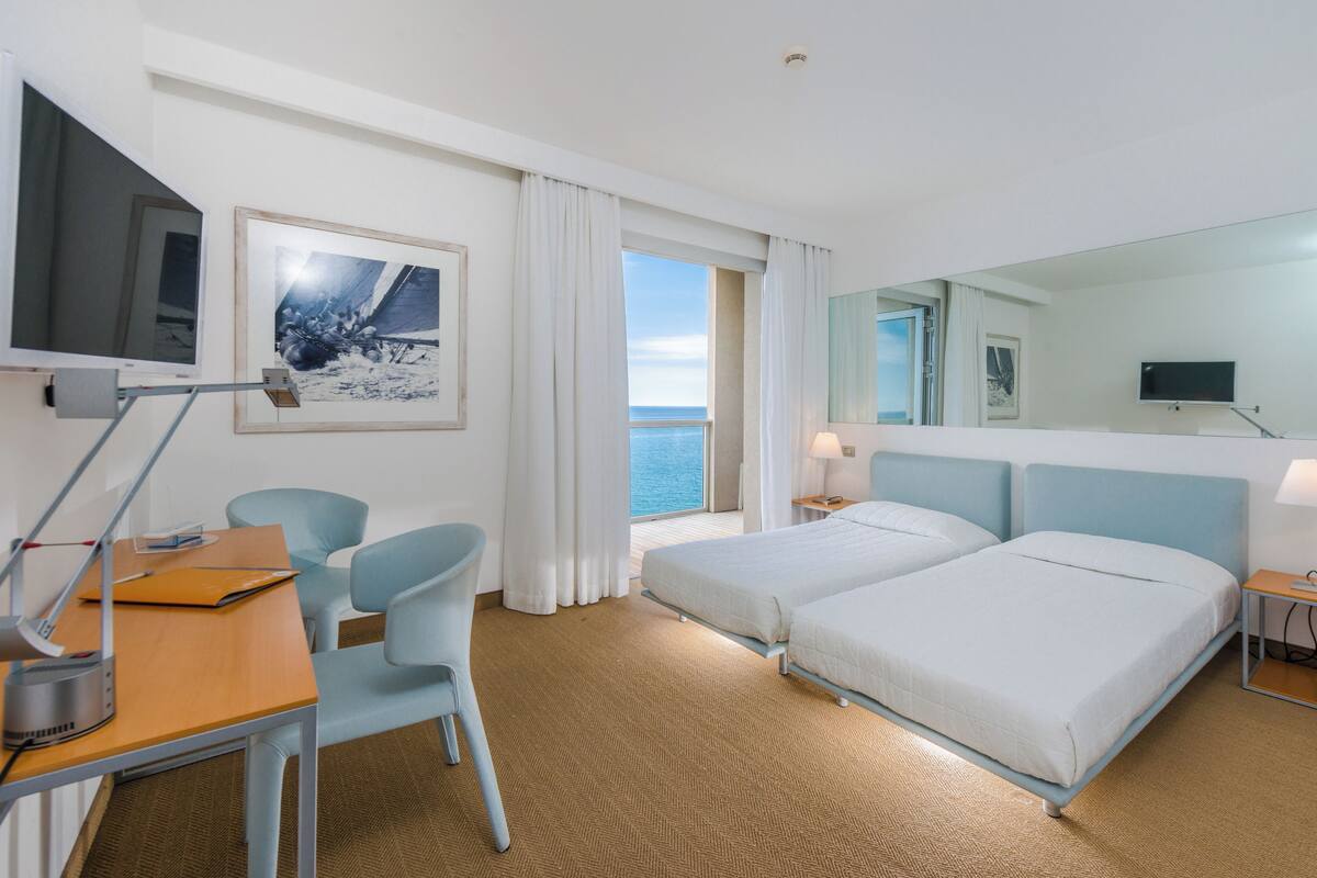 A hotel room with two beds, a desk with a chair, a TV, and a large window with a view of the sea.