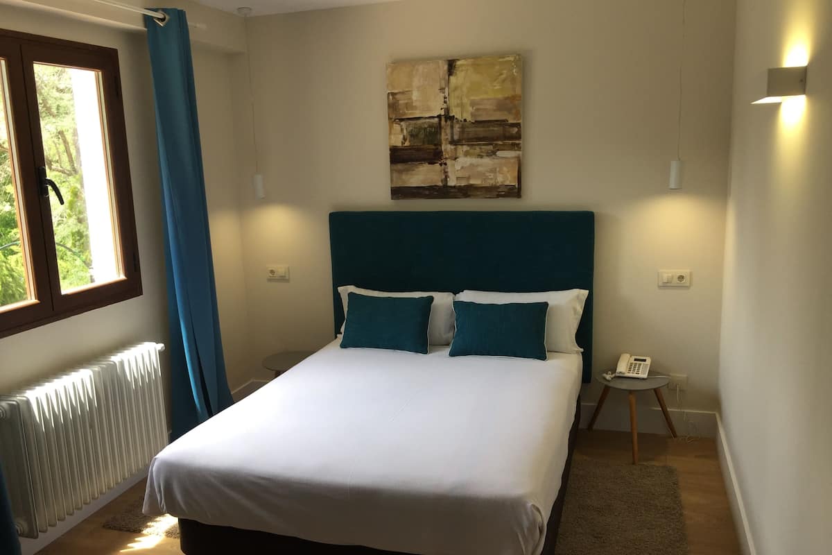 Un dormitorio con cama, ventana con cortinas, radiador, mesita de noche con teléfono y un cuadro en la pared.