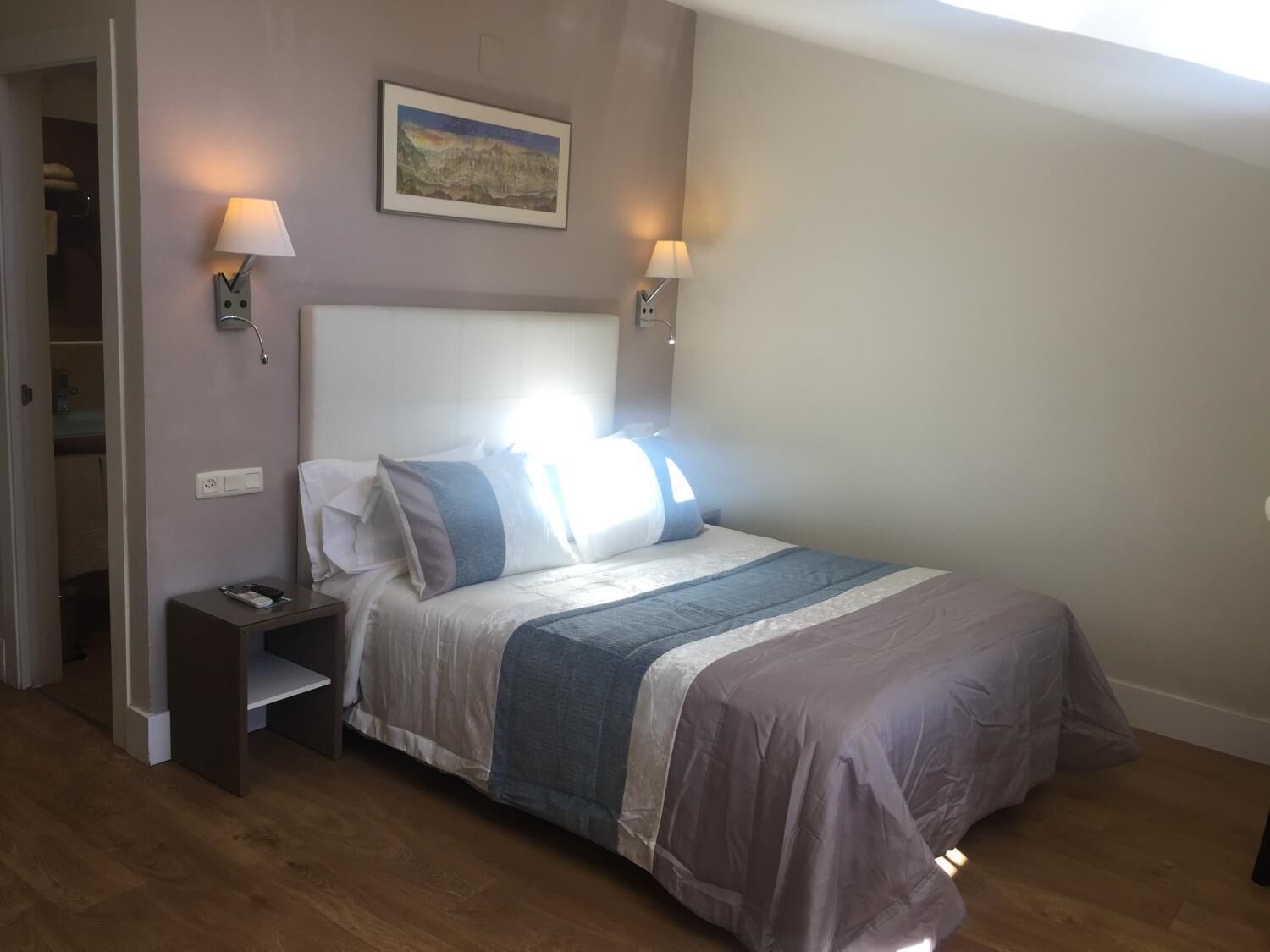 A bedroom with a bed, bedside lamps, a nightstand, a painting on the wall, and a door leading to a bathroom.
