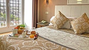 A neatly made bed with patterned pillows, a bedside table with a lamp, and a breakfast tray with fruits and a coffee cup. - Hotel Leonor de Aquitania (Cuenca)