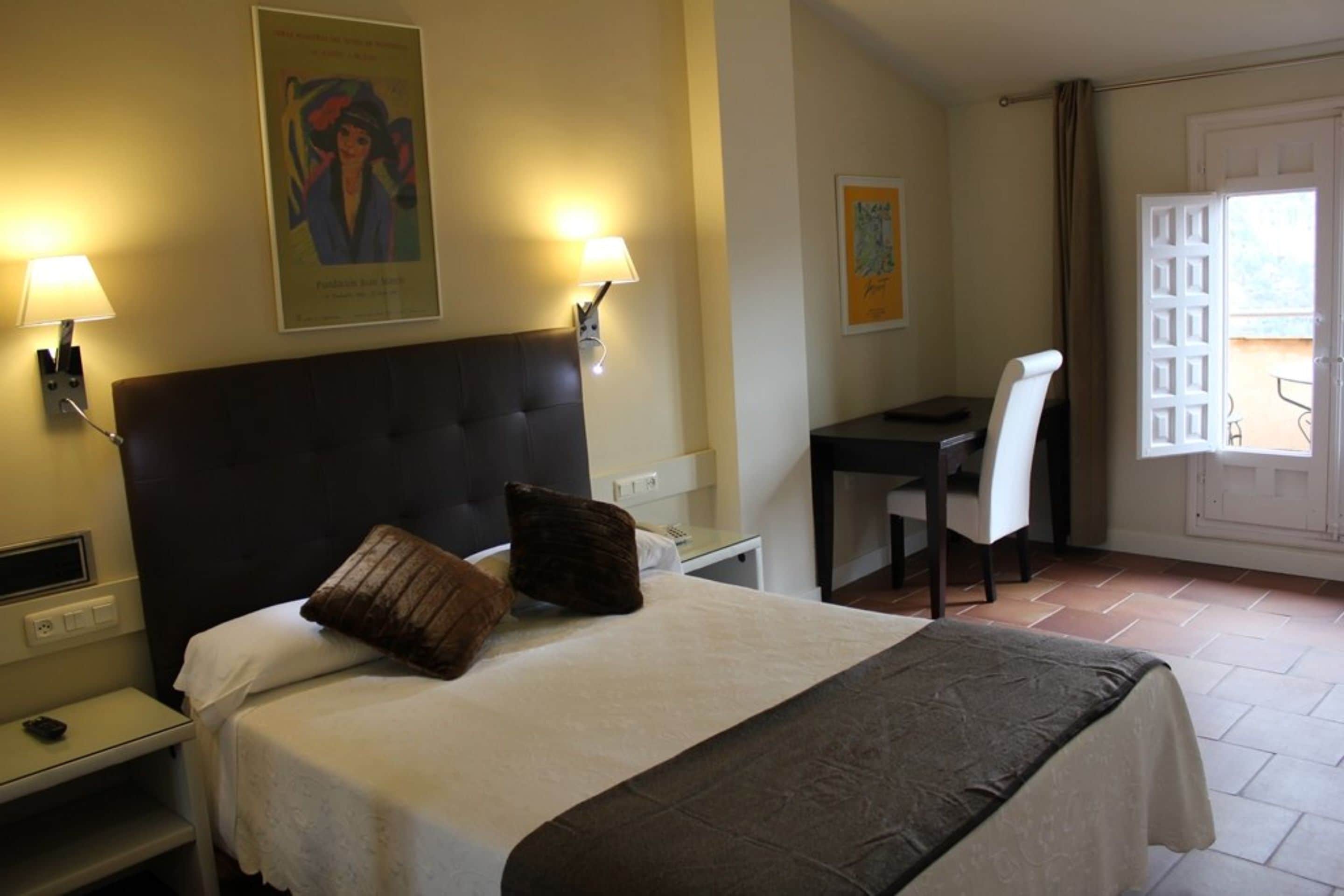 Een hotelkamer met een bed, nachtkastje, lamp en een bureau met stoel.