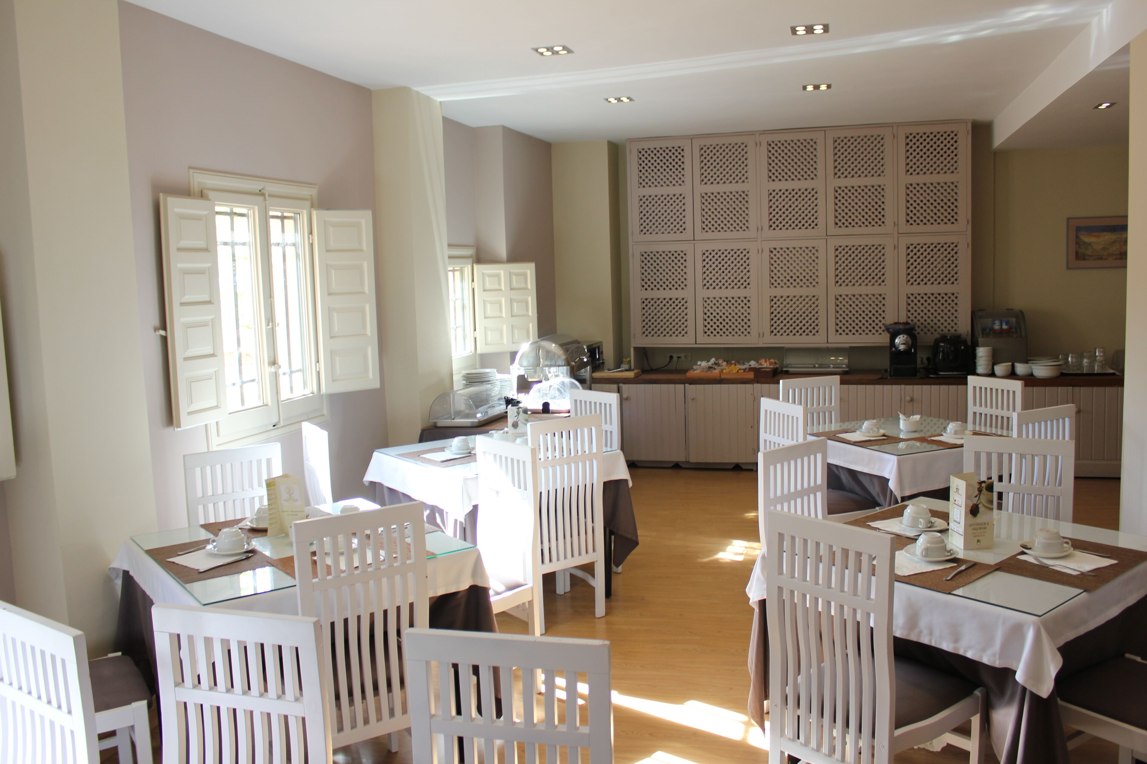 A dining area with white tables and chairs, a kitchen in the background, and a decorative wall panel.
