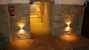 A stone-walled hallway with a fire extinguisher mounted on the left wall, a wooden door at the end, and two wall-mounted lights illuminating the path. - Hotel Leonor de Aquitania (Cuenca)