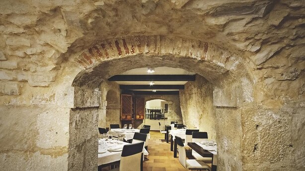 An interior with stone arches, tables set for dining, and a kitchen area in the background. - Hotel Leonor de Aquitania (Cuenca)