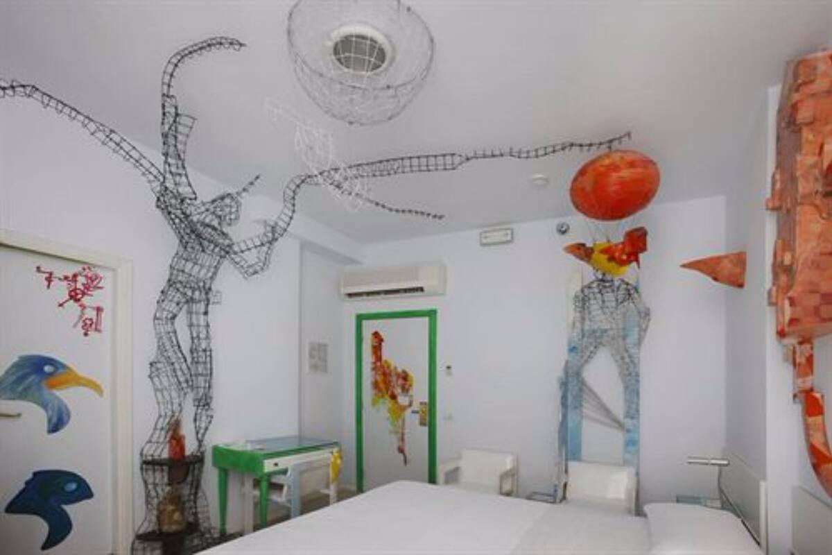 Ein Zimmer mit einem Bett, einem Schreibtisch und einem Stuhl. Eine Tür mit grünem Rahmen und eine Wand mit einer großen, abstrakten Drahtskulptur.