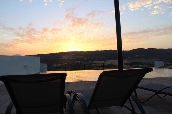 A terrace with two lounge chairs overlooking a body of water and distant hills during sunset.