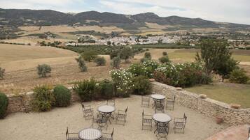 An outdoor patio with tables and chairs, surrounded by a stone wall and plants, overlooking a valley with fields and distant buildings.