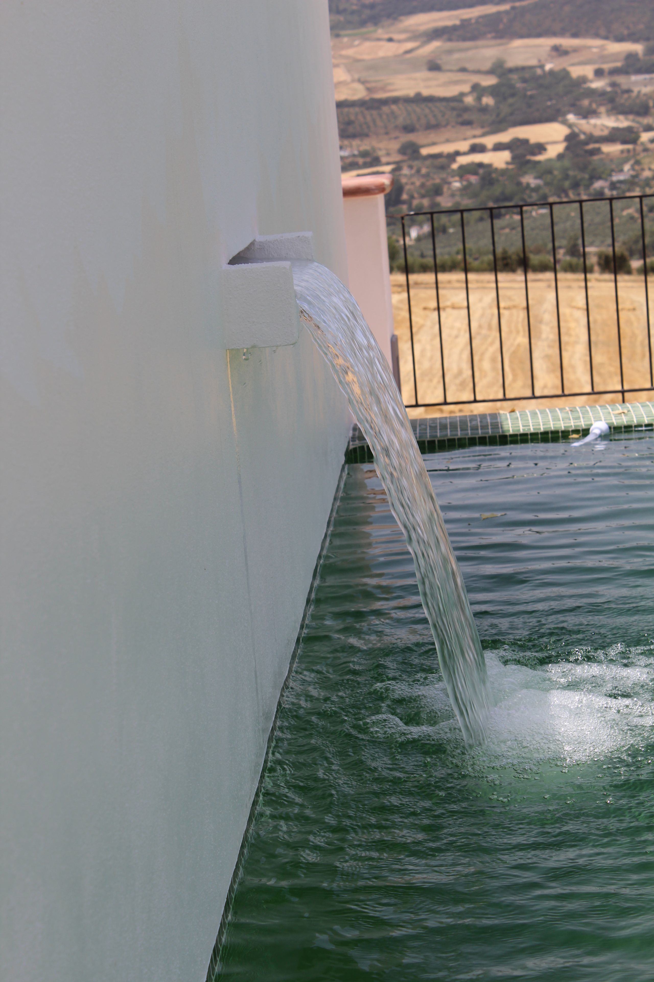 Una fuente de agua con un arroyo cristalino que desemboca en una piscina, rodeada por un muro blanco y una valla metálica.