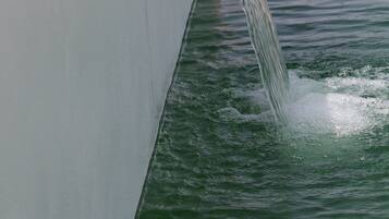 Una fuente de agua con un arroyo cristalino que desemboca en una piscina, rodeada por un muro blanco y una valla metálica.