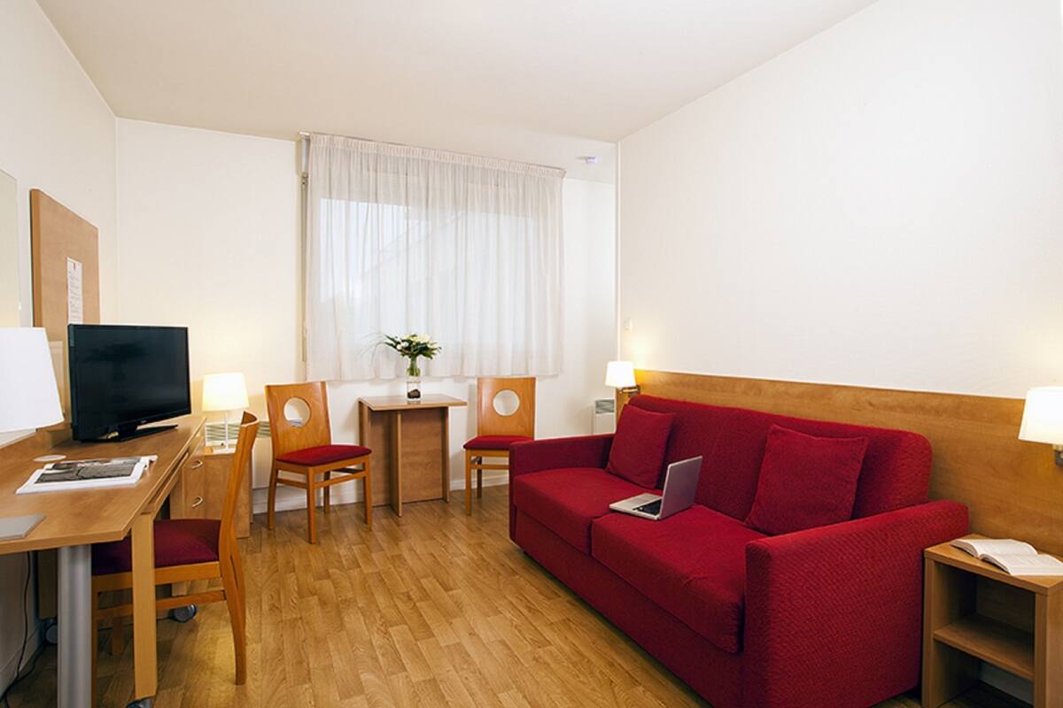 Habitación de hotel con un sofá rojo, un escritorio de madera con una computadora, un televisor y dos sillas.