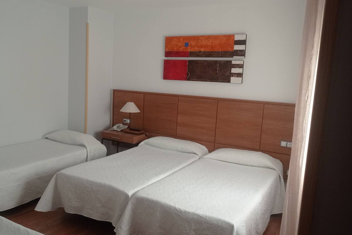 A hotel room with two single beds, a nightstand with a lamp, a telephone, and a wall-mounted artwork.