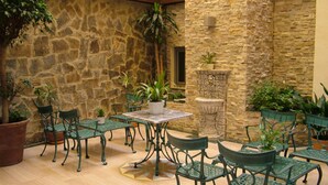 An outdoor patio with stone walls, green metal chairs, and tables, surrounded by potted plants.