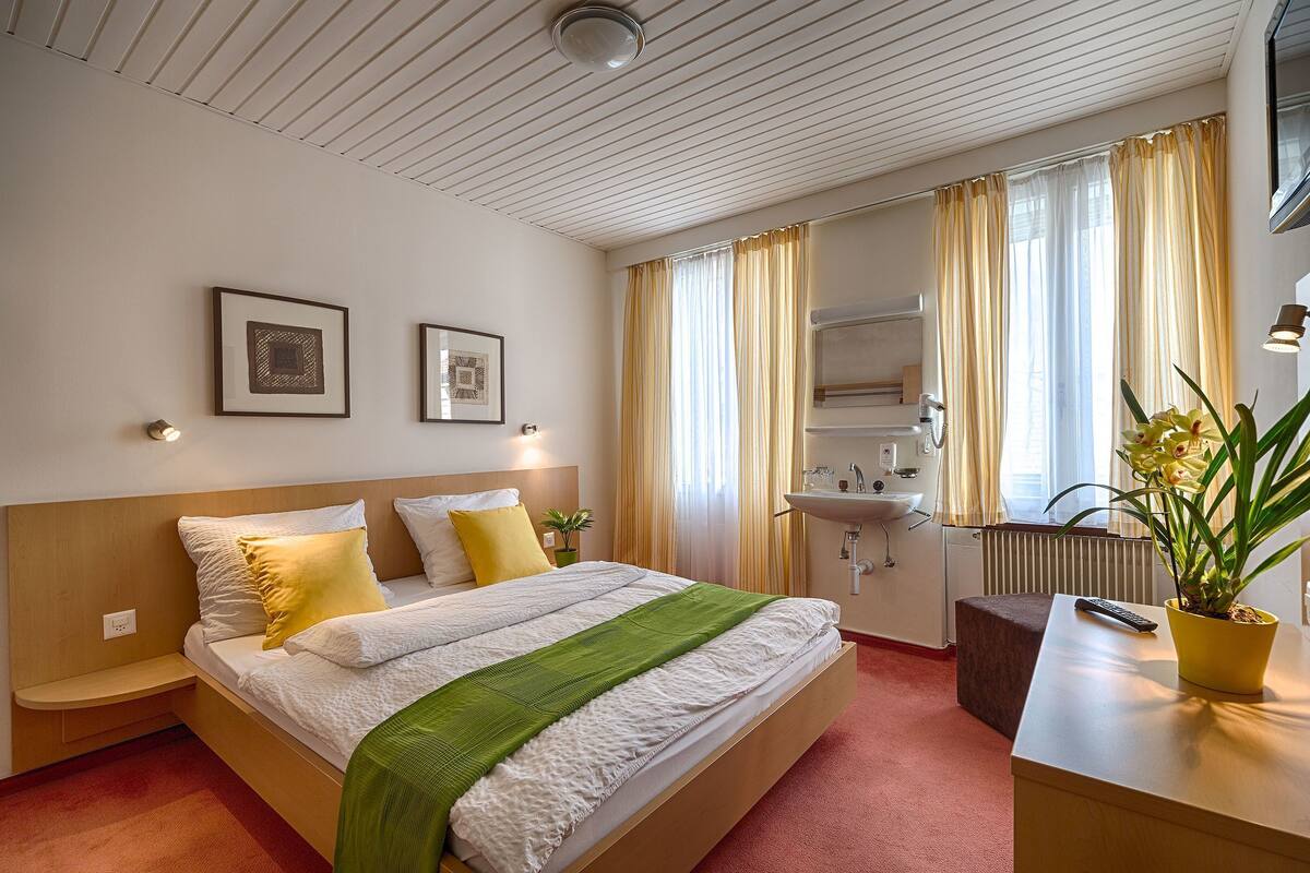 A hotel room with a bed, bedside table, potted plant, and a bathroom with a sink and mirror.