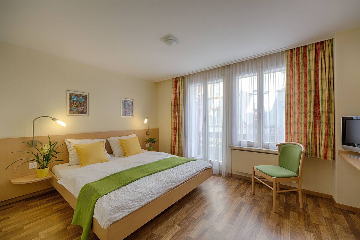 A hotel room with a bed, a chair, a TV, and a window with curtains.