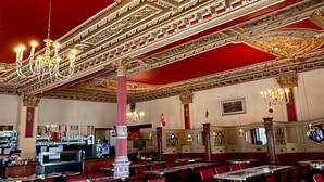 A grand dining hall with ornate ceiling decorations, red upholstered chairs, and marble tables. - Hotel Harder Minerva (Interlaken)
