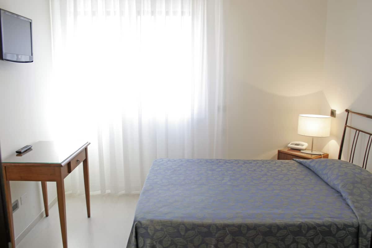 A hotel room with a bed, a desk, a chair, a nightstand with a lamp, and a telephone.