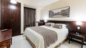 A hotel room with a large bed, a wooden wardrobe, a desk, and a painting of a cityscape.