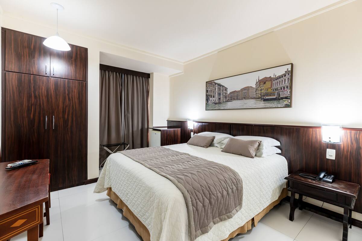 A hotel room with a large bed, a wooden wardrobe, a desk, and a painting of a cityscape.