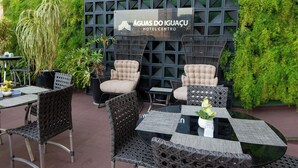 An outdoor seating area with wicker chairs and tables, a sign for ÁGUA DO IGUACU HOTEL CENTER, and lush greenery.