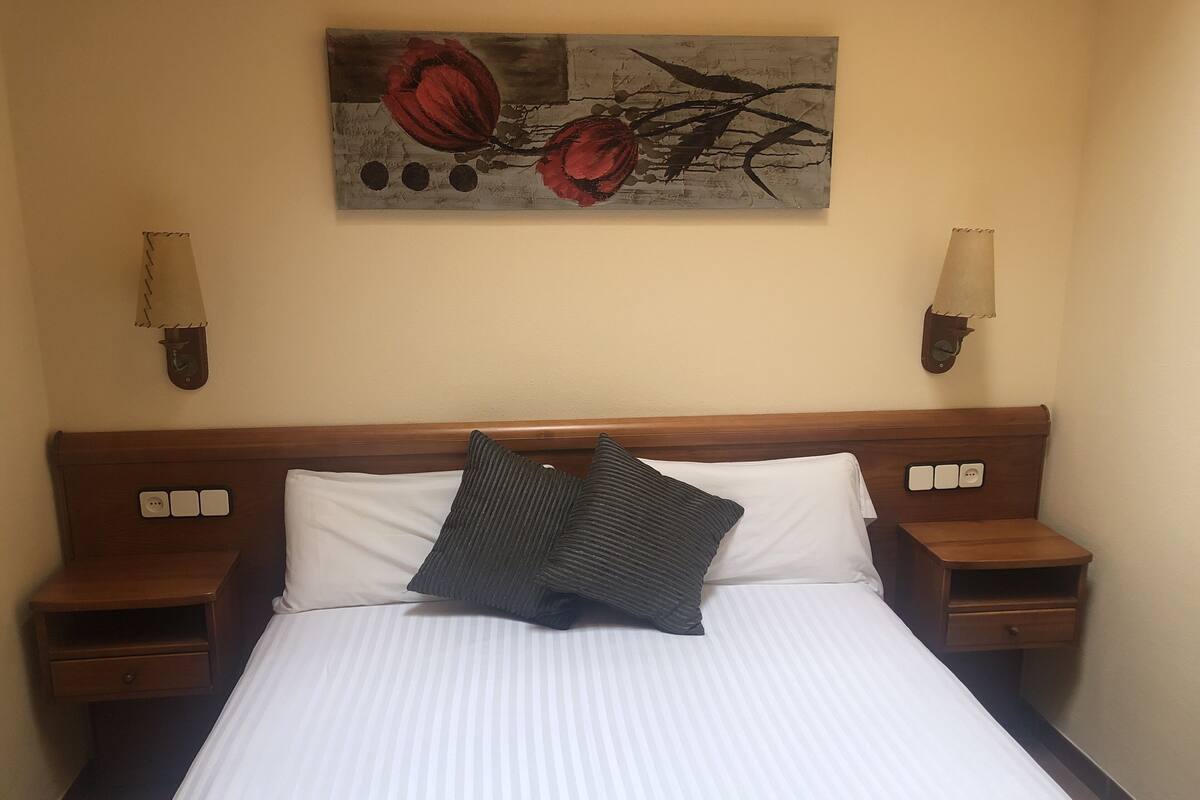 A hotel room with a large bed, two bedside tables, a nightstand, a wall-mounted lamp, and a painting above the bed.