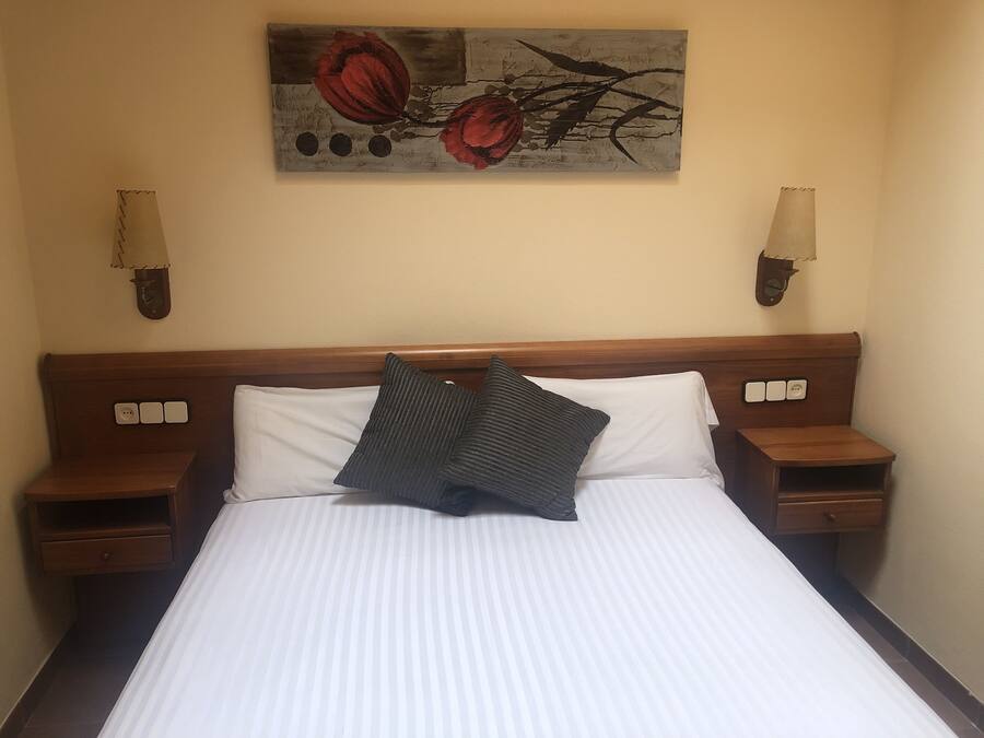 A hotel room with a large bed, two bedside tables, a nightstand, a wall-mounted lamp, and a painting above the bed.