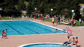 Una piscina pública amb diverses línies de bany, una piscina infantil i una zona enjardinada al voltant amb persones prenent el sol i passejant.