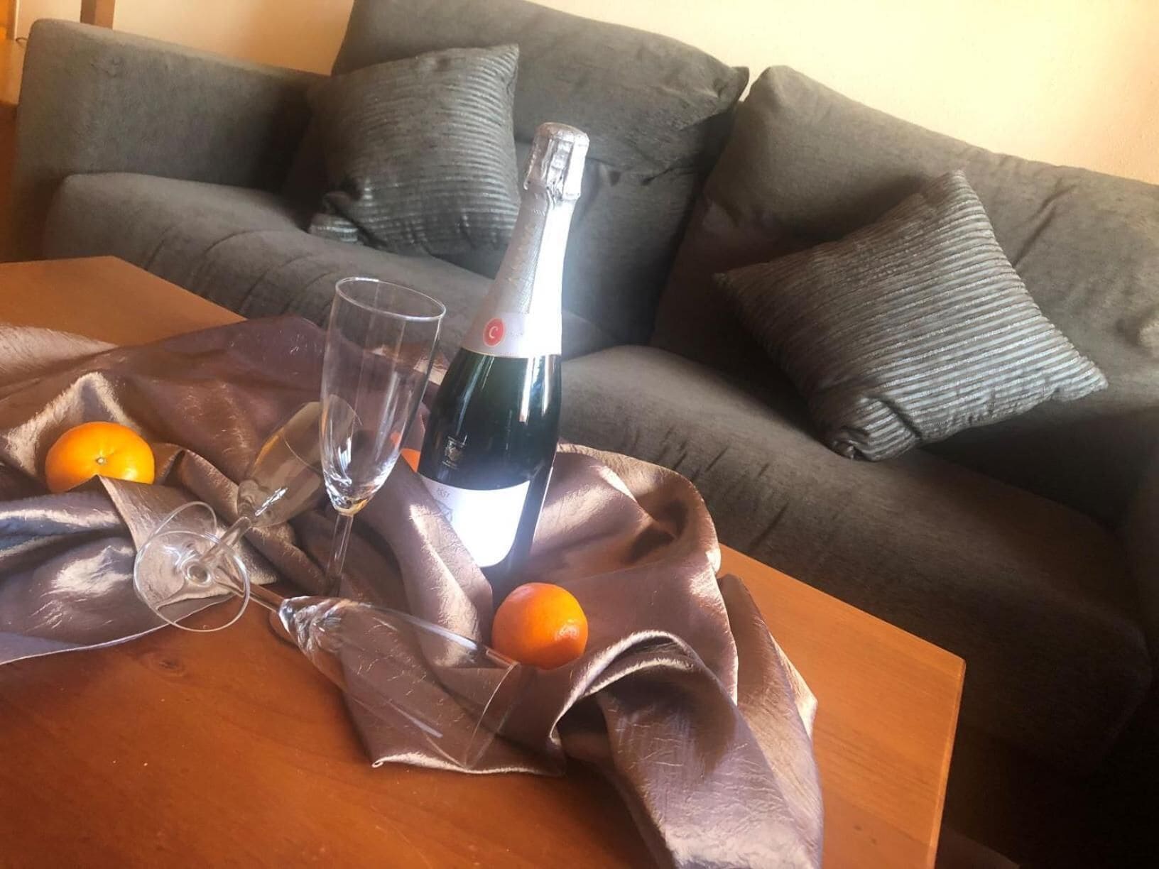 A table with a bottle of wine, two glasses, and two oranges on a tablecloth.