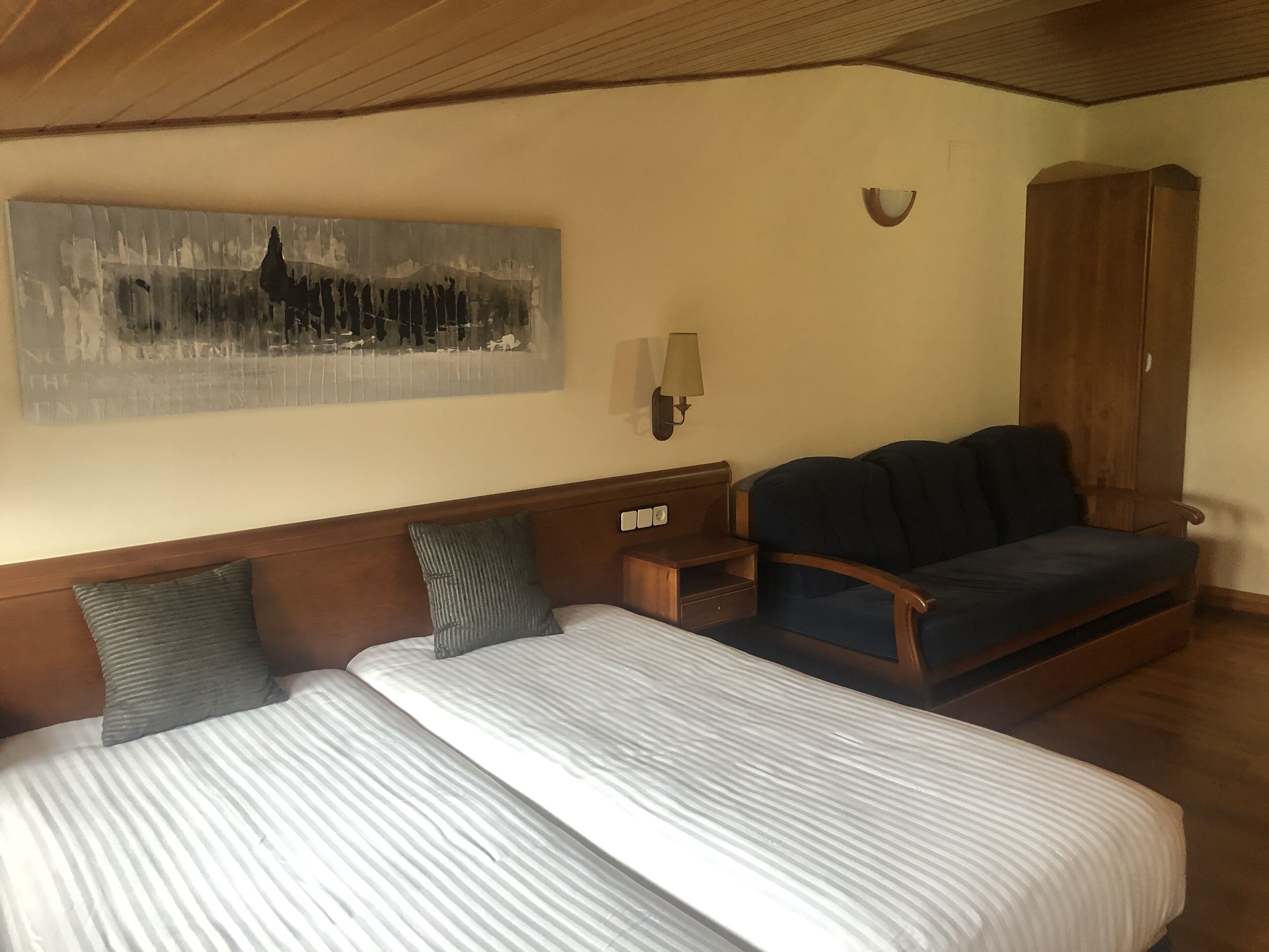 A hotel room with two beds, a wooden headboard, a dark sofa, a wooden wardrobe, and a wall-mounted lamp.