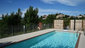 A rectangular swimming pool with a metal fence and a wooden fence, surrounded by greenery and houses in the background. - Villa Lauri (Neive)