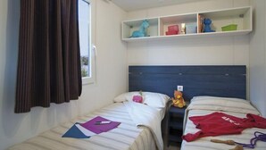 A bedroom with two beds, a window with curtains, and a shelf with toys and books. - Villaggio Camping Bosco Selva (Alberobello)