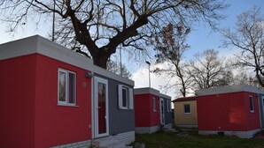 A row of small, single-story cabins with a red and gray exterior, a brick foundation, and a large tree in the background. - Villaggio Camping Bosco Selva (Alberobello)