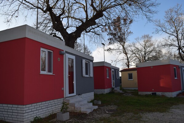 A row of small, single-story cabins with a red and gray exterior, a brick foundation, and a large tree in the background.