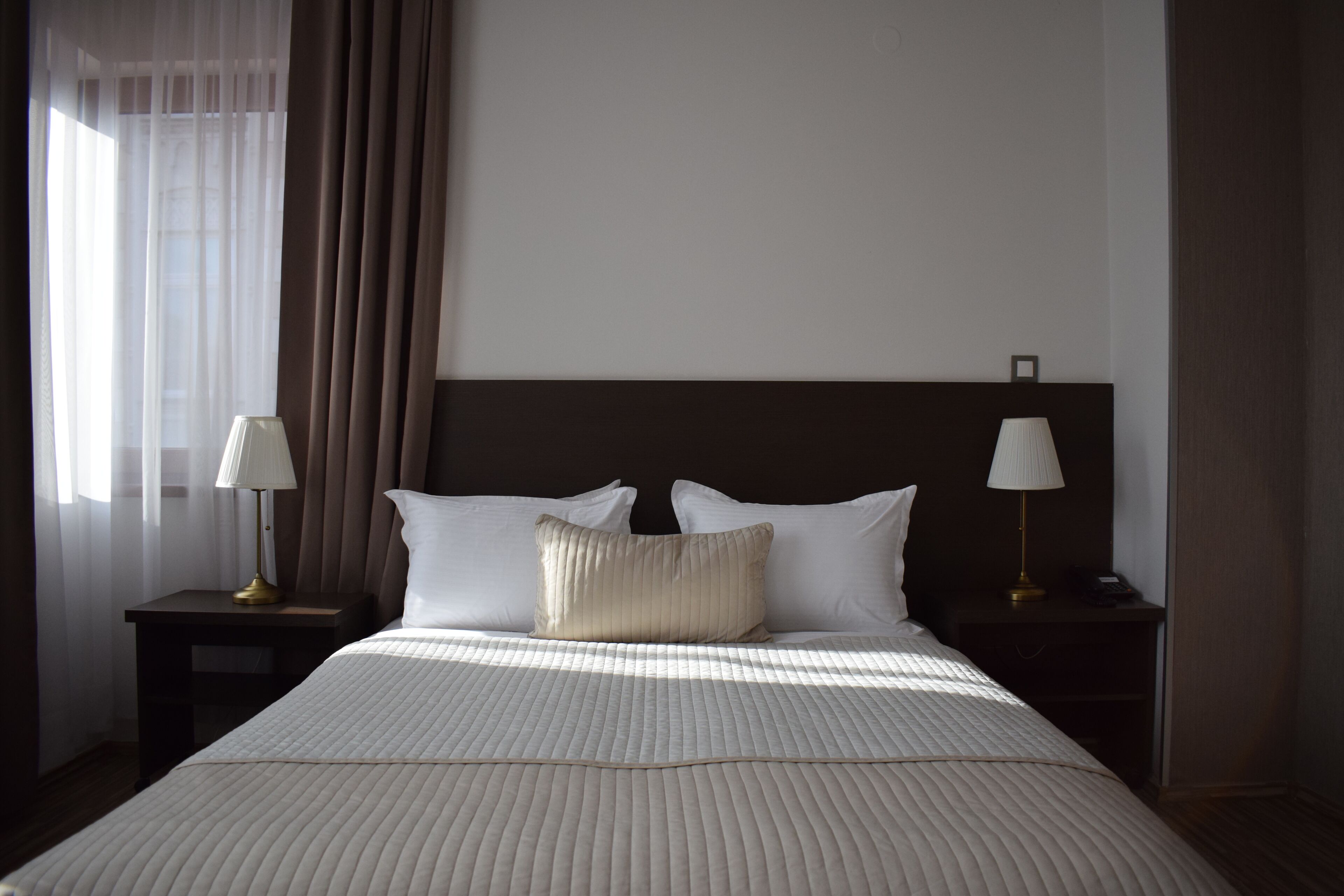 A hotel room with a large bed, two bedside tables with lamps, a headboard, and a window with curtains.