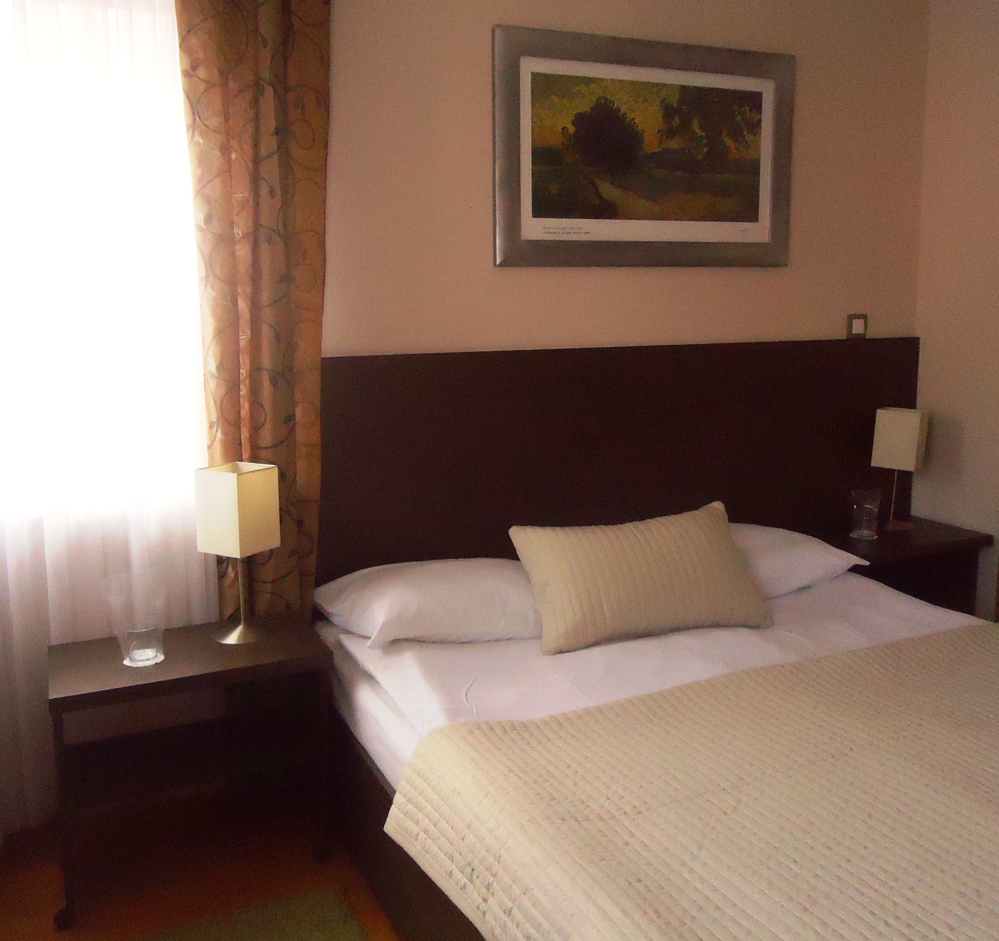 Une chambre d’hôtel avec un lit, une table de chevet, une lampe, un tableau au mur et une fenêtre avec des rideaux.