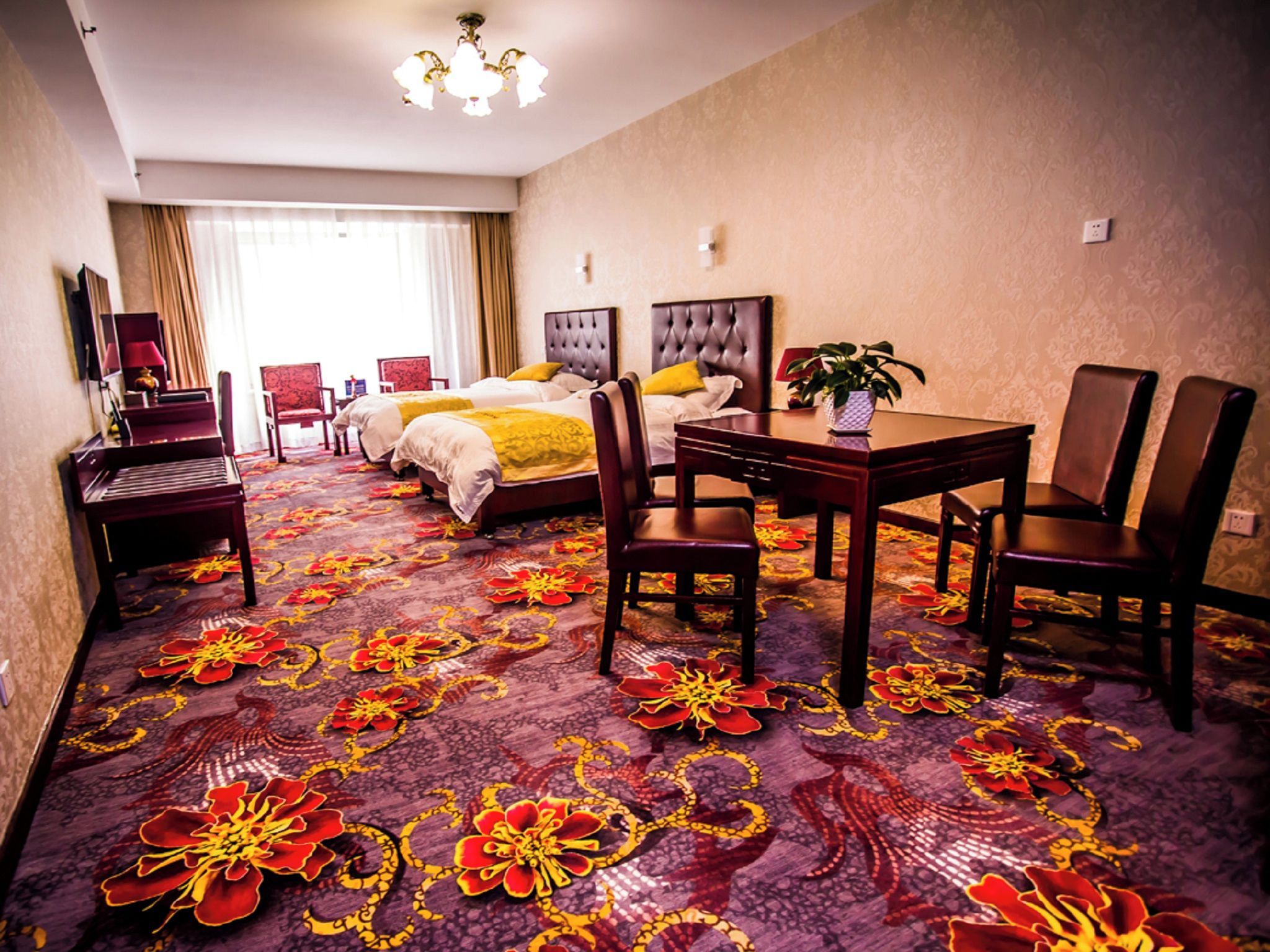 A hotel room with a large bed, two bedside tables, a desk with chairs, a potted plant, and a carpet with a floral pattern.