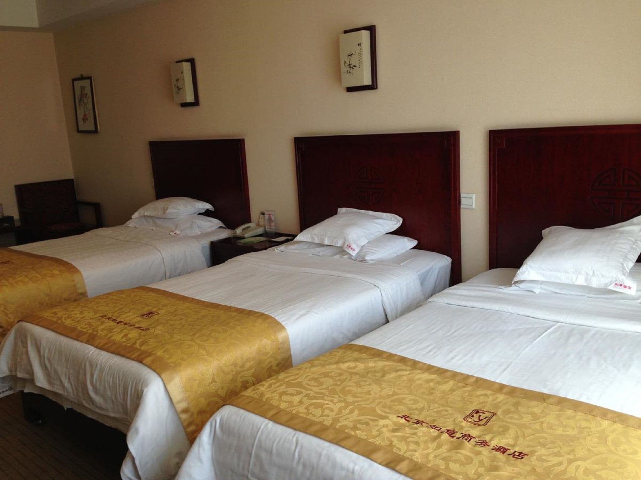 A hotel room with three single beds, each with a wooden headboard and white bedding. There are framed pictures on the wall and a bedside table with a lamp.