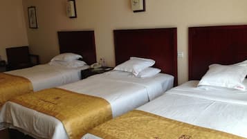A hotel room with three single beds, each with a wooden headboard and white bedding. There are framed pictures on the wall and a bedside table with a lamp.