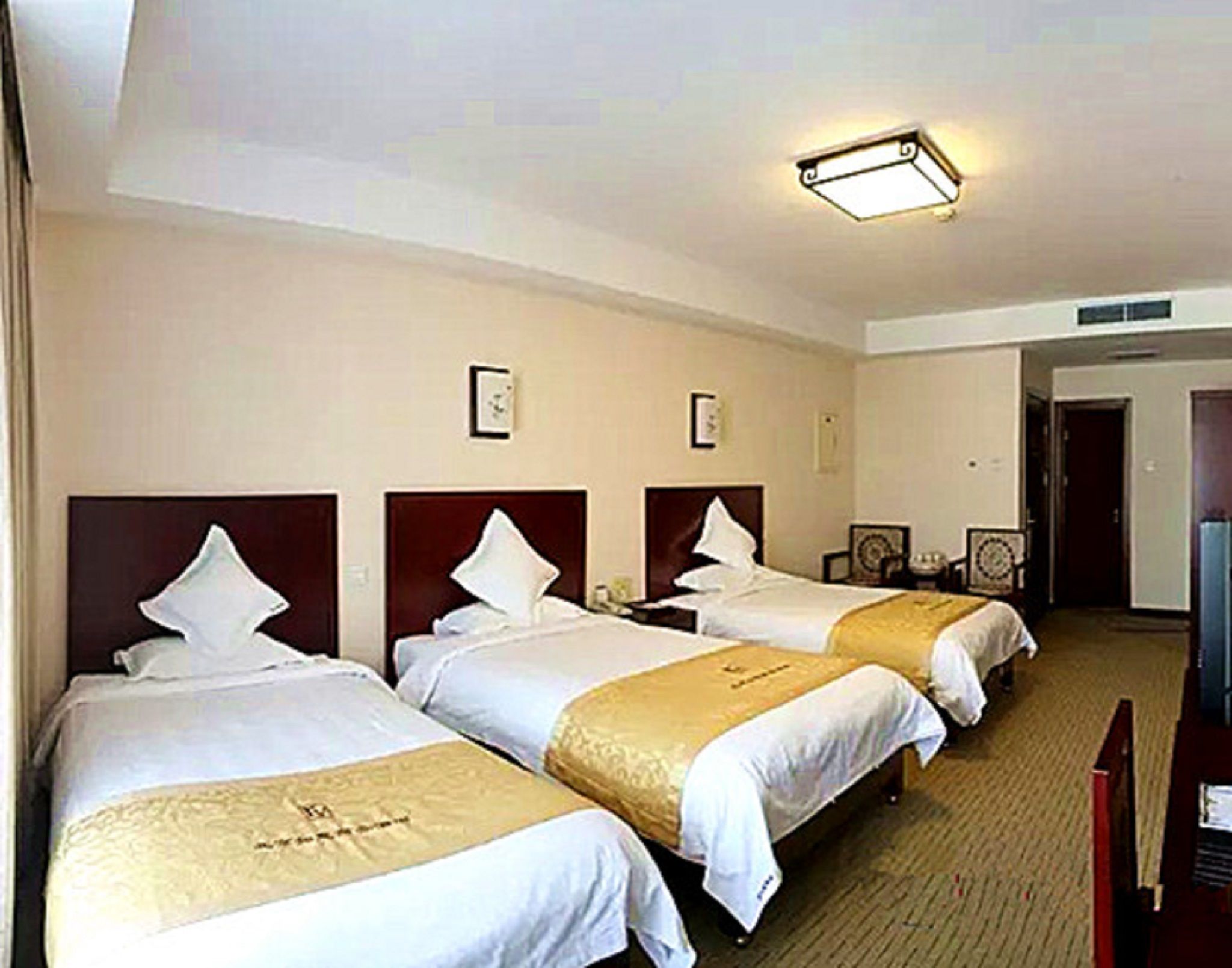 Ein Hotelzimmer mit drei Betten, jeweils mit Kopfteil und Zierkissen. Rechts daneben steht ein Fernseher auf einem Tisch und eine Tür führt in einen weiteren Raum.