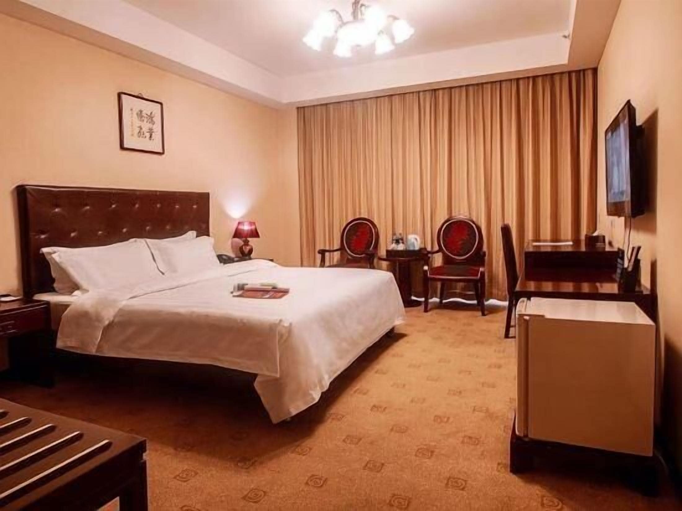 Quarto de hotel com uma cama, uma escrivaninha, duas cadeiras, uma televisão e uma pequena mesa com abajur.