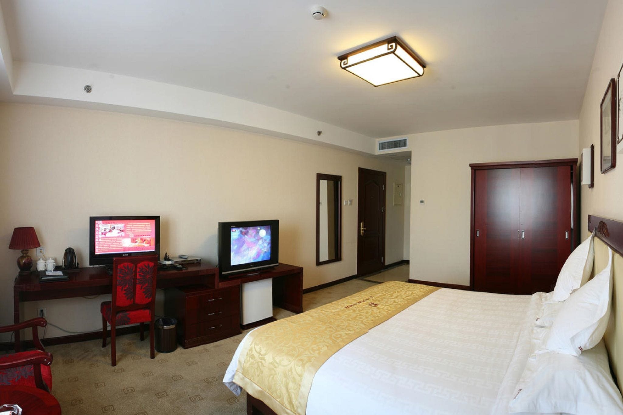 Ein Hotelzimmer mit einem Bett, zwei Fernsehern, einem Schreibtisch, einem Stuhl, einem Kleiderschrank und einem Spiegel.