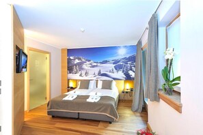Une chambre d’hôtel avec une grande fresque murale représentant un paysage de montagne enneigé, un lit recouvert d’une couette grise et blanche, et une petite orchidée en pot posée sur le rebord de la fenêtre.