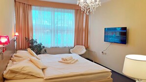 A hotel room with a bed, a TV, a chair, a lamp, and a window with curtains. - Hotel Casanova (Duchcov)