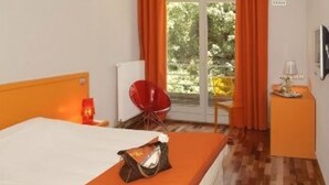 A hotel room with a bed, a red chair, a small table with a lamp, and a view of greenery through the window. - Hôtel du Lac (Guebwiller)